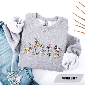 May include: Sport grey sweatshirt featuring a line of Disney characters: Pluto, Goofy, Donald Duck, Minnie Mouse, and Mickey Mouse. The sweatshirt has white lining at the cuffs and collar. The text "SPORT GREY" is at the bottom right.