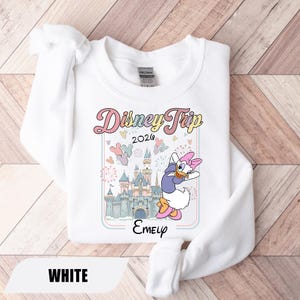 May include: White sweatshirt with a colourful graphic that reads "Disney Trip 2026" and a cartoon of Daisy Duck. The design includes a castle, balloons, and fireworks. The name "Emely" is printed below the graphic.