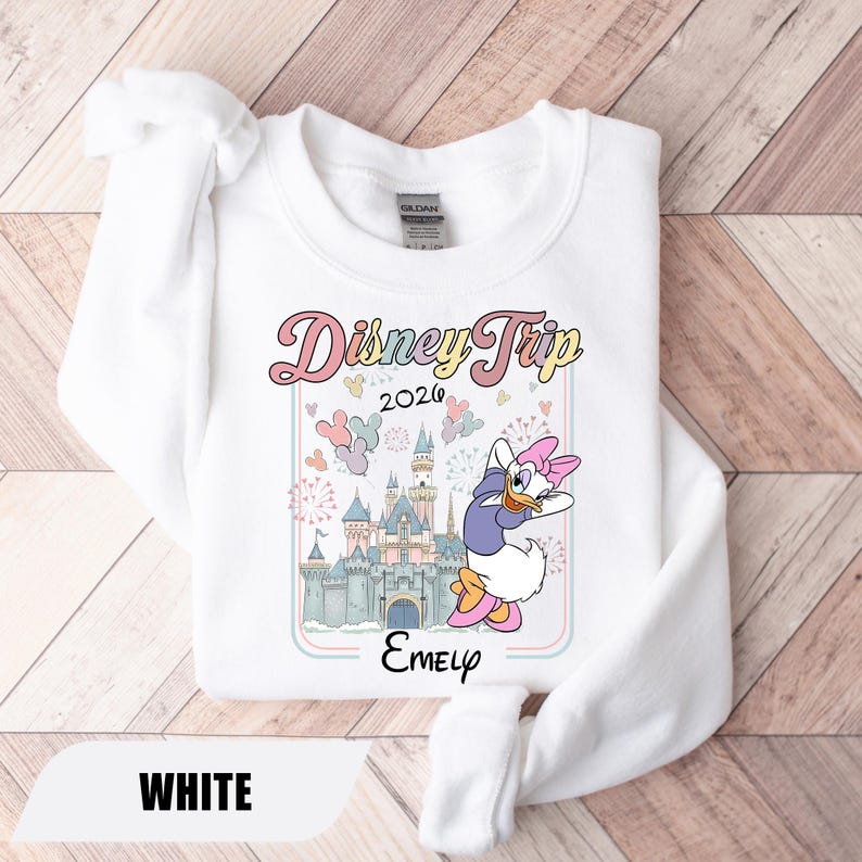 May include: White sweatshirt with a colourful graphic that reads "Disney Trip 2026" and a cartoon of Daisy Duck. The design includes a castle, balloons, and fireworks. The name "Emely" is printed below the graphic.