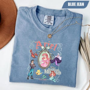 Puede incluir: Camiseta azul vaquero Comfort Colors con un gr&aacute;fico de Ariel de La Sirenita. El gr&aacute;fico incluye a Ariel, el Pr&iacute;ncipe Eric, Flounder, Sebasti&aacute;n y &Uacute;rsula. Tambi&eacute;n aparece el texto "Ariel" y "The Little Mermaid".