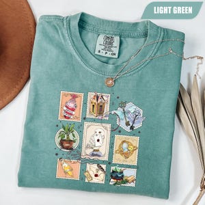 May include: Light green Comfort Colors t-shirt with a graphic of fantasy-themed postage stamps. A silver necklace with a pendant rests on the shirt. The shirt is folded, with the tag visible.