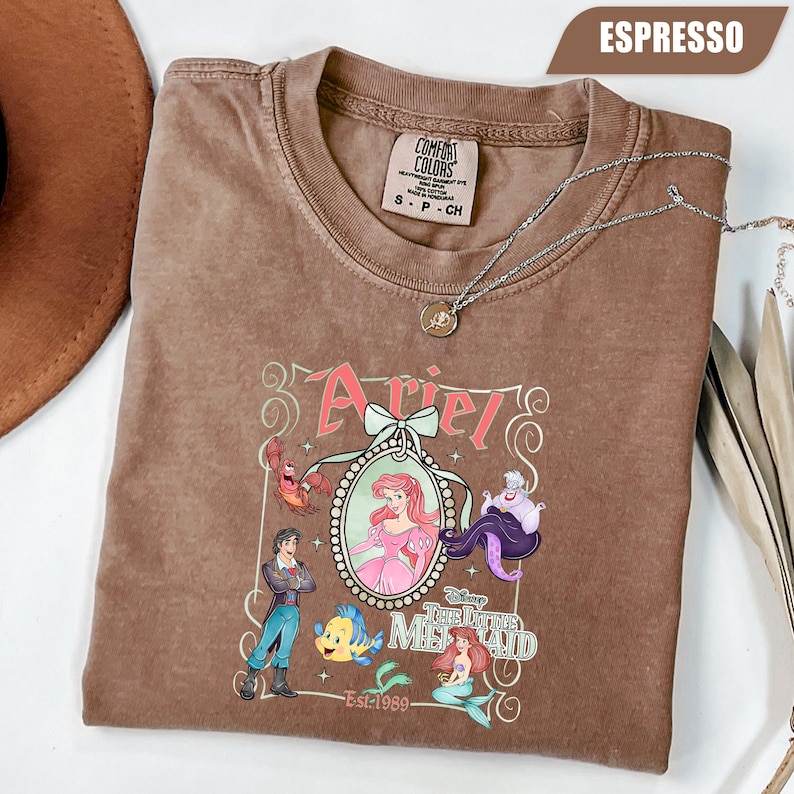 Puede incluir: Camiseta color espresso con un gr&aacute;fico de Ariel de La Sirenita, con personajes como el Pr&iacute;ncipe Eric, Flounder y &Uacute;rsula. El dise&ntilde;o incluye el texto "Ariel" y "La Sirenita" con el a&ntilde;o "Est. 1989". Un collar plateado est&aacute; drapeado sobre la camiseta.