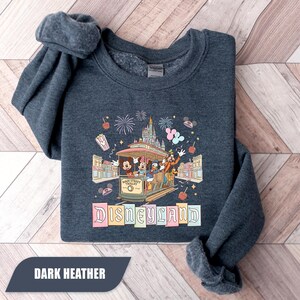 Pode incluir: Camisola cinza escuro com um gr&aacute;fico colorido da Disneyland. O design mostra Mickey, Minnie, Pato Donald e Pateta em um bonde da Main Street. Fogos de artif&iacute;cio, um castelo e a palavra "Disneyland" tamb&eacute;m s&atilde;o exibidos.