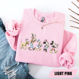 May include: Light pink sweatshirt featuring a graphic of Disney characters: Pluto, Goofy, Donald Duck, Minnie Mouse, and Mickey Mouse. The characters are walking and holding food items. The text "LIGHT PINK" is at the bottom of the image.