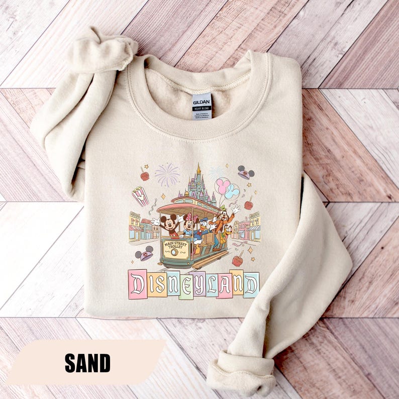Pode incluir: Camisola cor de areia com um gr&aacute;fico da Disneyland. O design inclui um bonde da Main Street com Mickey Mouse, Minnie, Pato Donald e Pateta, juntamente com um castelo, bal&otilde;es e fogos de artif&iacute;cio. A palavra "Disneyland" est&aacute; escrita abaixo.