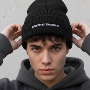 May include: Black ribbed knit beanie with the text "ENCRYPTED THOUGHTS" embroidered in white. The beanie is worn low on the forehead. The person in the image is wearing a gray hooded sweatshirt.