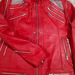 May include: A vibrant red leather jacket featuring a stand-up collar, multiple silver-toned metal zips across the chest and sleeves, and decorative silver rhinestone panels on both shoulders. The front includes a central zip closure.