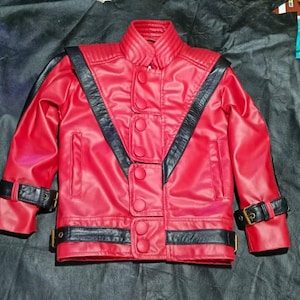 May include: A bright red jacket featuring black leather-like accents, a high collar, and a front closure with multiple circular buttons. The garment includes black straps with gold-toned buckles on the cuffs and hem, and ribbed shoulder detailing.