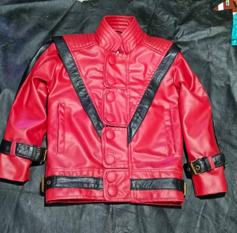 May include: A bright red jacket featuring black leather-like accents, a high collar, and a front closure with multiple circular buttons. The garment includes black straps with gold-toned buckles on the cuffs and hem, and ribbed shoulder detailing.