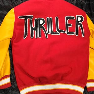 May include: A red varsity jacket with yellow sleeves and a white and yellow striped collar, cuffs, and hem. The back features the word THRILLER in large, black, white-outlined letters. The jacket has a snap-button front closure and a red body.