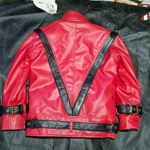 May include: A bright red jacket featuring prominent black leather-like V-shaped panels on the back. The garment includes black buckled straps at the waist and on each sleeve cuff, with a high collar and a smooth, structured finish throughout.