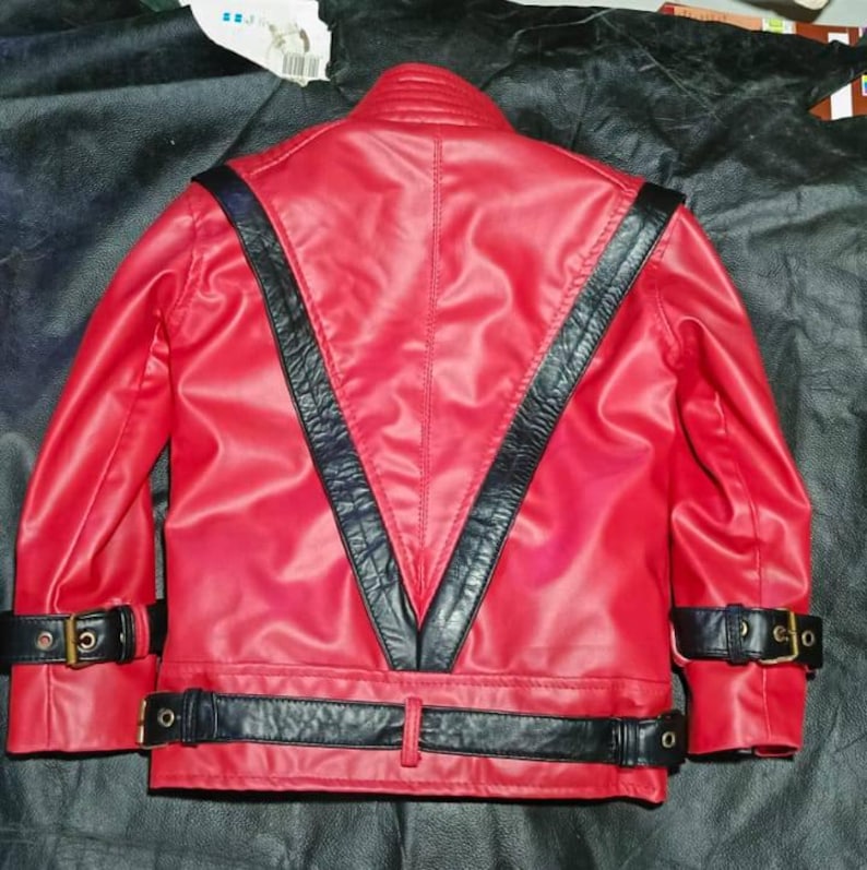 May include: A bright red jacket featuring prominent black leather-like V-shaped panels on the back. The garment includes black buckled straps at the waist and on each sleeve cuff, with a high collar and a smooth, structured finish throughout.