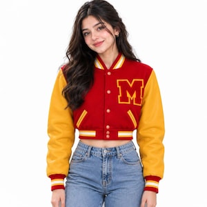 Pode incluir: Uma jaqueta varsity curta com corpo de lã vermelha e mangas amarelas. A frente apresenta um grande emblema com a letra M amarela no peito, fecho de botões e punhos e bainha canelados com listras brancas e amarelas.