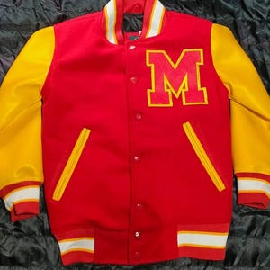May include: A red varsity jacket with yellow sleeves, featuring a large red letter M with a yellow border on the left chest. The jacket has a snap-button front closure, two yellow-trimmed pockets, and striped ribbed cuffs and hem in red and white.