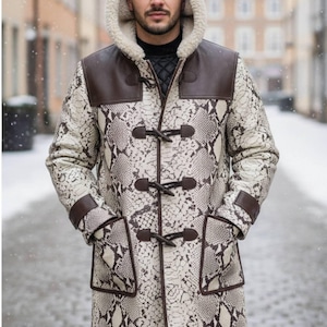 May include: A long coat with a snakeskin pattern in cream and brown, featuring a hood with a cream-colored lining. The coat has brown leather accents on the shoulders, cuffs, and pockets, and is fastened with wooden toggles. The coat is worn in a snowy outdoor setting.
