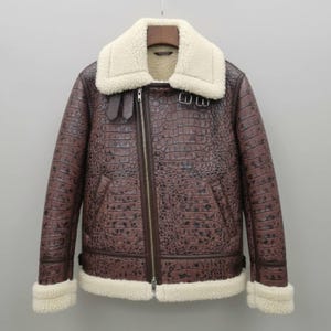 Könnte beinhalten: Braune Lederjacke mit Schafsfellkragen und -futter. Die Jacke hat eine Krokodilhautstruktur, einen Reißverschluss und Schnallendetails. Die Manschetten und der Kragen sind mit cremefarbenem Schafsfell gefüttert.
