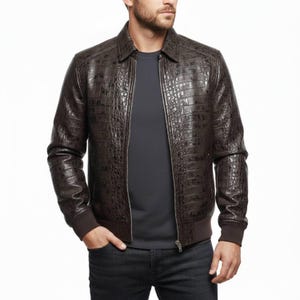 Puede incluir: Chaqueta de cuero marrón con textura de piel de cocodrilo. La chaqueta tiene cierre de cremallera, cuello y puños y dobladillo de canalé. Se lleva sobre una camiseta gris oscuro y vaqueros oscuros.