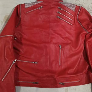 May include: A bright red jacket featuring silver-toned hardware, including multiple zipped pockets and accents. The shoulders are embellished with panels of sparkling clear rhinestones. The jacket has a high collar and long sleeves with zips.