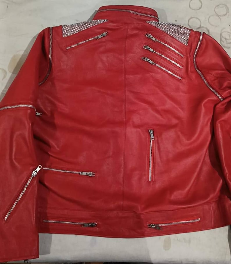 May include: A bright red jacket featuring silver-toned hardware, including multiple zipped pockets and accents. The shoulders are embellished with panels of sparkling clear rhinestones. The jacket has a high collar and long sleeves with zips.