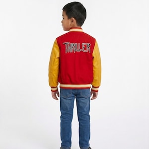 May include: A red and yellow varsity jacket featuring the word THRILLER embroidered on the back in a stylised font. The jacket has long yellow sleeves, a red body, and striped ribbed trim at the collar, cuffs, and hem, paired with blue denim jeans.