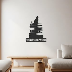 Puede incluir: Arte de pared de metal negro que representa un gato sentado sobre una pila de libros. Las palabras "CUSTOM NAME" se muestran en la parte inferior. Esta obra de arte está diseñada para la decoración del hogar y añade un toque literario.