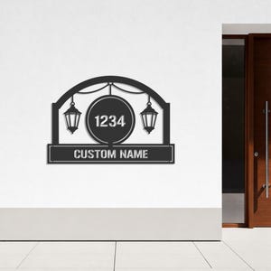 Pode incluir: Placa de casa em metal preto com um topo arqueado decorativo, apresentando dois desenhos de lanternas e um círculo central com o número 1234. A placa inclui o texto "CUSTOM NAME" em uma base retangular.