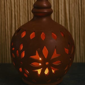 以下が含まれることがあります： 切り抜かれた花と葉の模様が施された茶色のセラミックランタン。球形の本体は、装飾的な上部を備えた球根状の形状をしています。ろうそくの温かいオレンジ色の光が切り抜きを通して輝き、居心地の良い雰囲気を作り出しています。