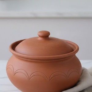 Puede incluir: Una olla de arcilla de color terracota con tapa, de forma redondeada y con patrones decorativos grabados. La olla está sobre una superficie de mármol, junto a una cuchara de madera y un paño de lino. Un artículo de cocina.
