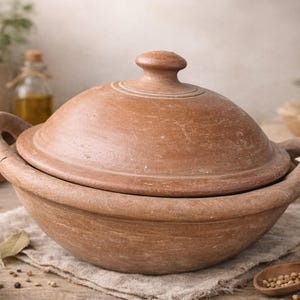 Może przedstawiać: Terakotowy garnek do gotowania z pokrywką, dwoma uchwytami i gałką. Garnek stoi na lnianej szmatce, otoczony przyprawami i ziołami. Garnek ma średnicę około 25 cm.