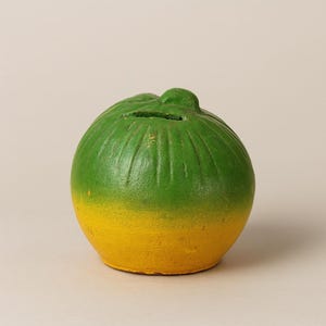 Könnte beinhalten: Eine Keramik-Spardose in Form einer Frucht, mit einem grünen Oberteil und einem gelben Unterteil. Die Spardose hat einen Schlitz für Münzen und eine strukturierte Oberfläche. Ein dekorativer Artikel zum Sparen von Geld.