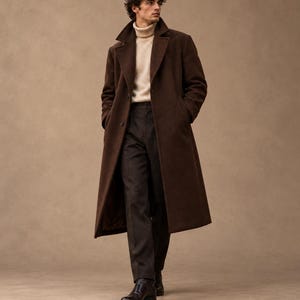 Pode incluir: Um casaco comprido de lã castanho-escuro com lapelas largas e botões, usado sobre uma camisola de gola alta canelada em tom creme e calças cinzento-carvão. O visual é completado com botas de couro castanho-escuro.