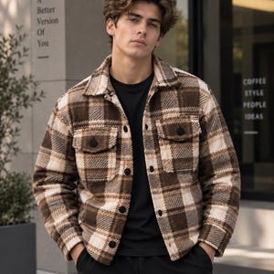 Könnte beinhalten: Eine braun-beige karierte Hemdjacke mit Kragen, langen Ärmeln und zwei Brusttaschen mit schwarzen Knöpfen. Das Kleidungsstück wird über einem schlichten schwarzen T-Shirt und einer schwarzen Hose getragen. Der Stoff hat eine dicke, strukturierte Optik.