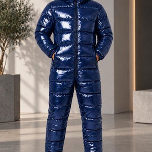 May include: A glossy navy blue puffer jumpsuit featuring a full-length front zip, a hood, and horizontal quilted channels. The garment has long sleeves and full-length trouser legs with elasticated cuffs, paired with clean white athletic trainers.
