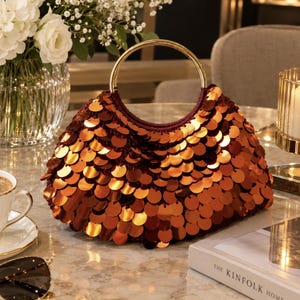 May include: A handbag covered in overlapping, reflective copper-colored circular sequins. The bag features a dark maroon fabric base and a single, polished gold-tone circular metal handle attached to the top, creating a structured, rounded silhouette.