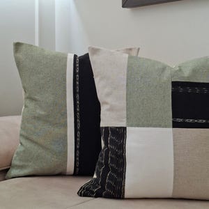 May include: Two decorative throw pillows with a patchwork design. The pillows feature a mix of green, black, white, and beige fabric. One pillow has a vertical stripe pattern, while the other has a geometric block pattern. The pillows are on a beige sofa.