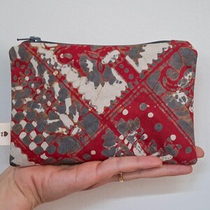 May include: A rectangular pouch with a red, white, and grey patterned design. The pouch has a zip closure and a small tag on the side. The pouch is held in a person's hand.