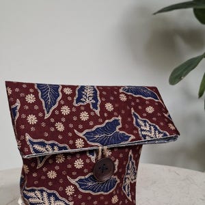 May include: A maroon and navy blue batik fabric pouch with a button closure. The pouch features a floral and leaf pattern in navy blue and cream against a maroon background. The button is brown and the pouch is sitting on a white surface.