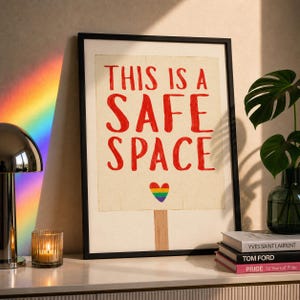 May include: A framed art print with the words "THIS IS A SAFE SPACE" in red. A rainbow heart is below the text. The print is in a black frame and is displayed on a white surface with books and a candle.