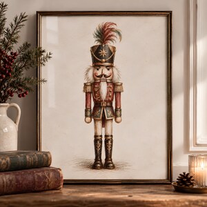 May include: A framed wall art print featuring a detailed illustration of a nutcracker soldier. The figure wears a black hat with a multi-coloured feather plume, a red jacket with gold trim, and black boots. The background is a neutral off-white.