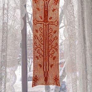 May include: A rectangular, rust-colored, crocheted wall hanging with a central cross design and floral accents. The piece is suspended in front of a window with white lace curtains. The window overlooks a snowy outdoor scene.