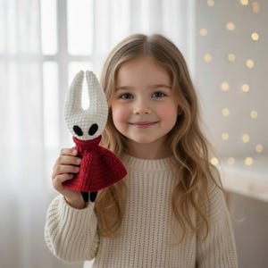May include: A handmade crochet doll featuring a white head with two horns, black eyes, and a red dress. The doll has black legs and is held up by a person wearing a cream-colored sweater. The background is a soft focus with a window and lights.