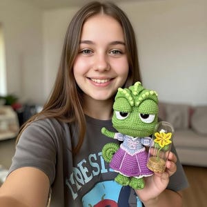Puede incluir: Una muñeca de camaleón verde de ganchillo hecha a mano con un vestido morado y un pequeño recipiente de vidrio con una flor amarilla. La muñeca tiene un diseño detallado y es sostenida por una persona.