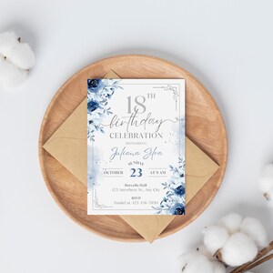May include: An invitation card for an 18th birthday celebration, featuring a floral design in shades of blue. The card includes the name "Juliana Silva", date, time, and location details. It rests on a wooden plate with a brown envelope.