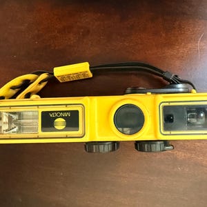 May include: A bright yellow Minolta Weathermatic 35mm film camera featuring a rectangular body, a built-in flash, a black lens cover, and a black wrist strap. The camera has black control dials on the bottom and a yellow strap attachment.