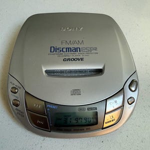 Könnte beinhalten: Ein silberner Sony Discman tragbarer CD-Player mit FM/AM-Radio. Das Gerät verfügt über ein digitales LCD-Display, das die Radiofrequenz 97.90 anzeigt, sowie über Bedientasten für Wiedergabe, Pause, Sendersuche und Titelnavigation und Beschriftungen wie ESP2, Groove und D-F411.