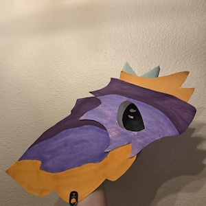 May include: A handcrafted paper mask featuring a stylized creature design with a pointed orange snout, a purple face section, and a light blue crown-like detail at the top. The mask includes a single dark eye opening and a black metal ring attachment.