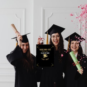 Puede incluir: Tres graduadas con togas y birretes negros celebran. Una sostiene un diploma, otra una tarjeta que dice "SHE DID IT! The future is yours. CLASS OF 2026" con detalles dorados. Confeti rosa cae a su alrededor.