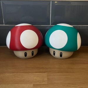 May include: Two mushroom-shaped figures, one with a red cap and one with a teal cap, both featuring white circular spots on the top. Each figure has a beige base with two black oval eyes printed on the front, creating a distinct character design.