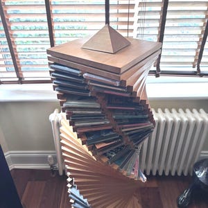 Puede incluir: Una torre de almacenamiento de CD de madera con un diseño en espiral. La torre está hecha de madera de color marrón claro y presenta una pirámide en la parte superior. La torre está llena de CD y se encuentra en un piso de madera cerca de una ventana con persianas.