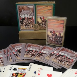 May include: A deck of playing cards featuring hunting scenes on the backs, displayed with a matching box. The cards show traditional face card designs, a joker, and numbered cards including a three of hearts, all spread out on a flat surface.